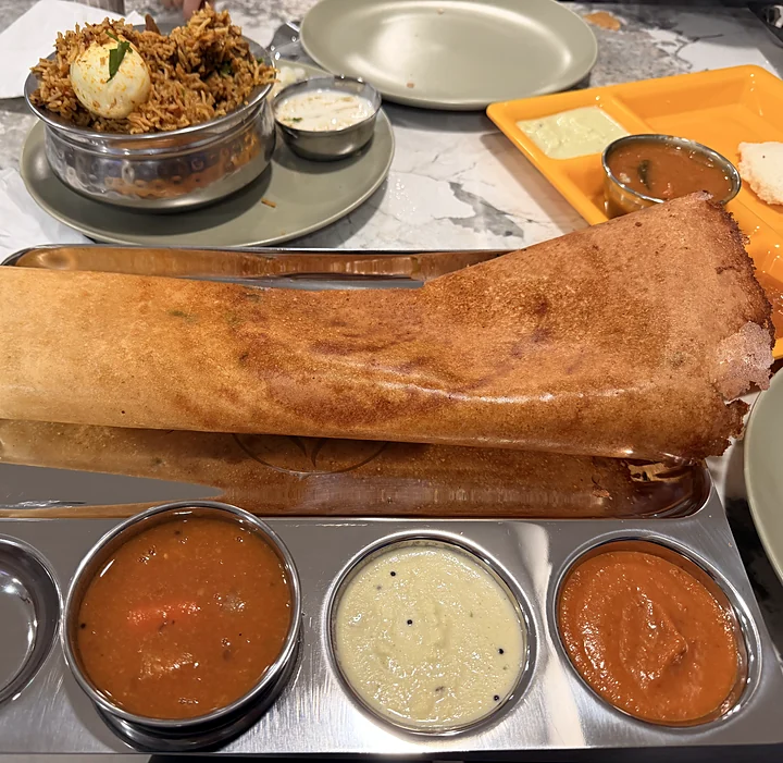Dosa on a steel thali with sambar, coconut chutney and tomato chutney, with biryani and idly on the table behind