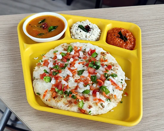 Mixed vegetable uttappam topped with onion, tomato, carrot and capsicum, with sambar and chutneys on a yellow tray